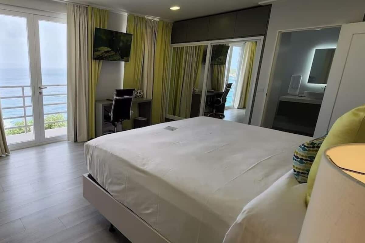 Habitación de hotel con una cama grande, un escritorio, una silla y un televisor. Tiene un balcón con vistas al océano.