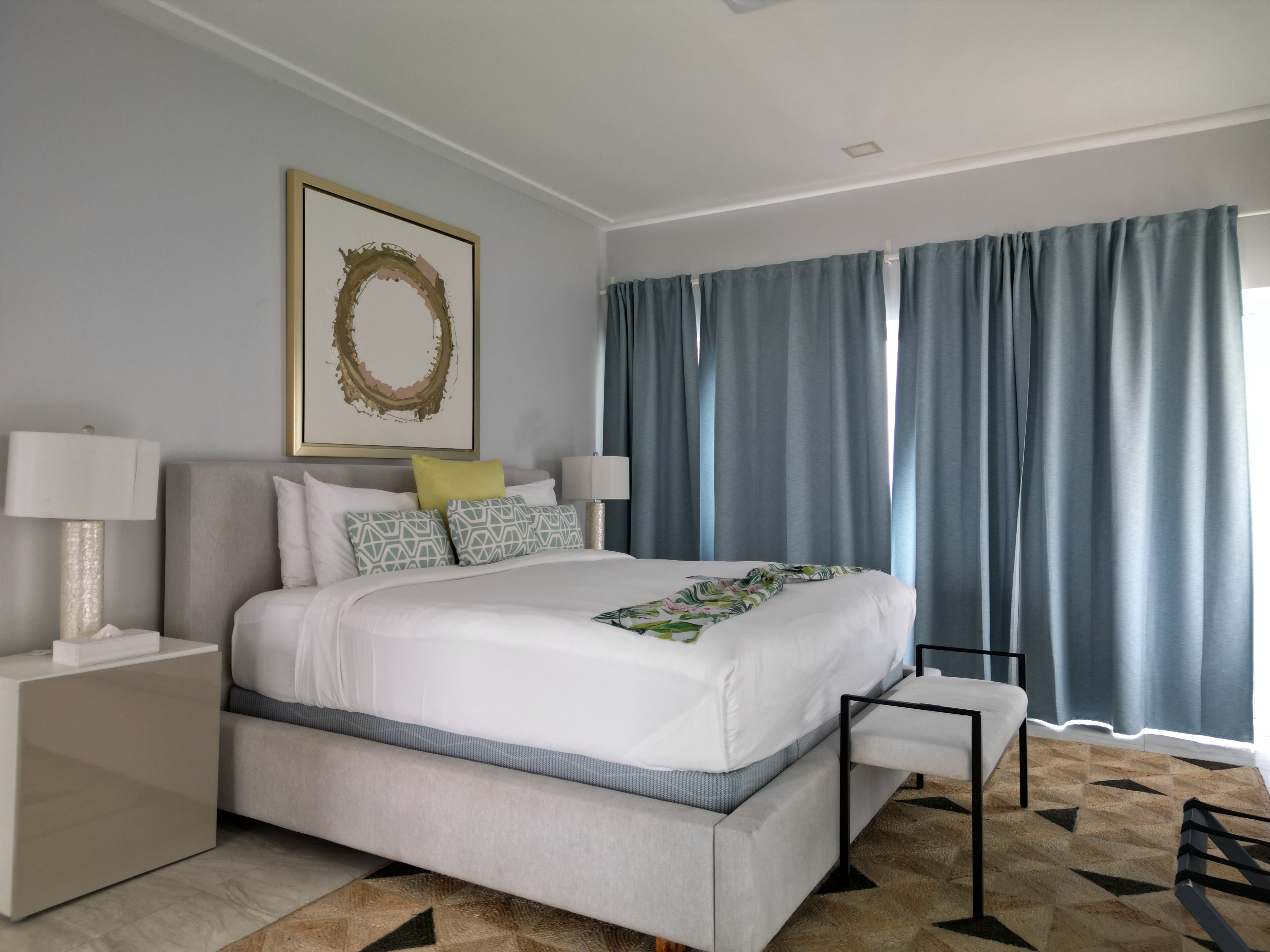 Een moderne hotelkamer met een groot bed, een nachtkastje met een lamp, een decoratief kunstwerk aan de muur en uitzicht door dunne gordijnen.