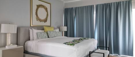 Een moderne hotelkamer met een groot bed, een nachtkastje met een lamp, een decoratief kunstwerk aan de muur en uitzicht door dunne gordijnen.