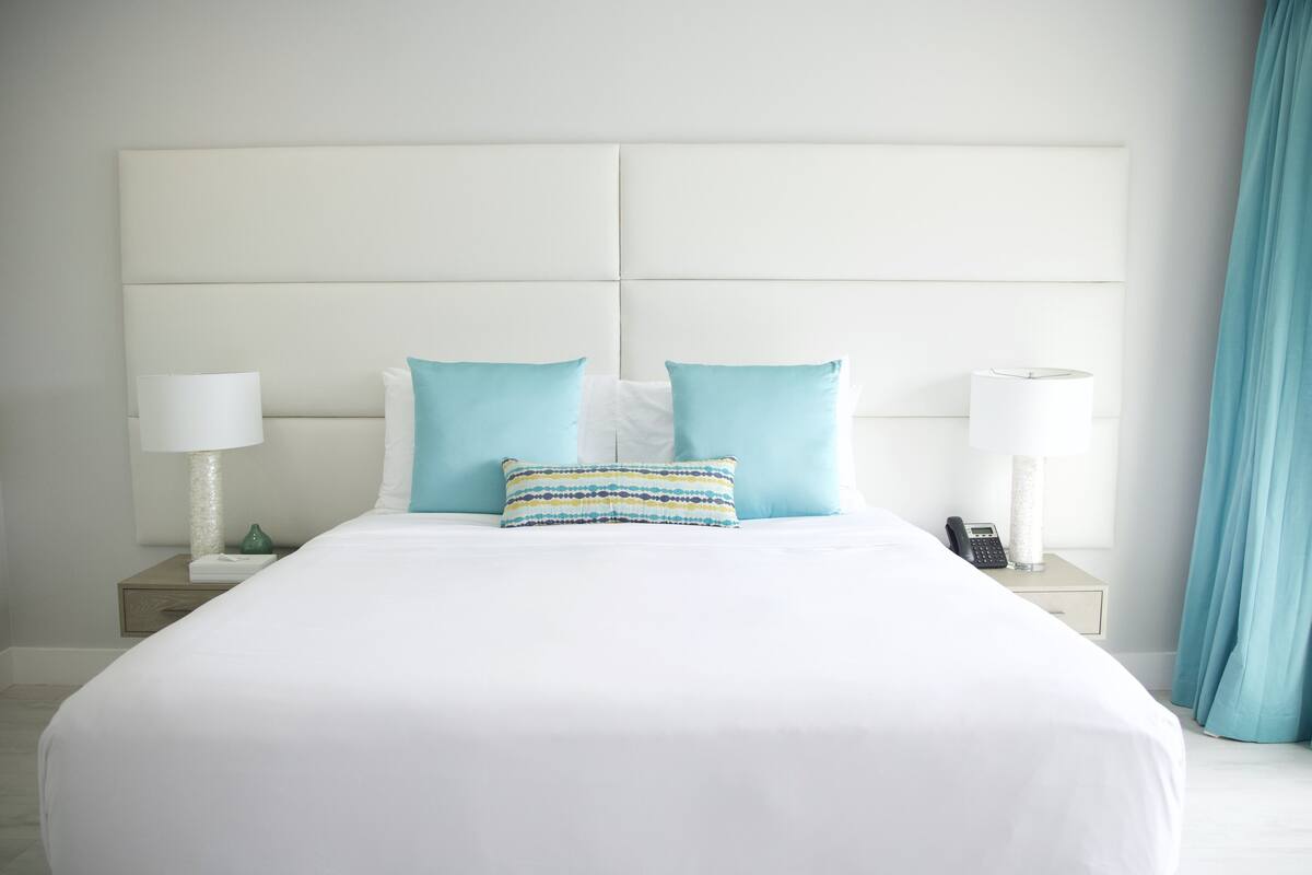 Una cama bien tendida con ropa blanca y dos almohadas azul claro, flanqueada por mesitas de noche con lámparas y un teléfono.