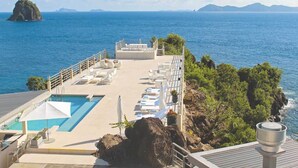 Zona junto a la piscina con sillones de descanso y sombrillas, con vista a un mar azul y una costa rocosa.