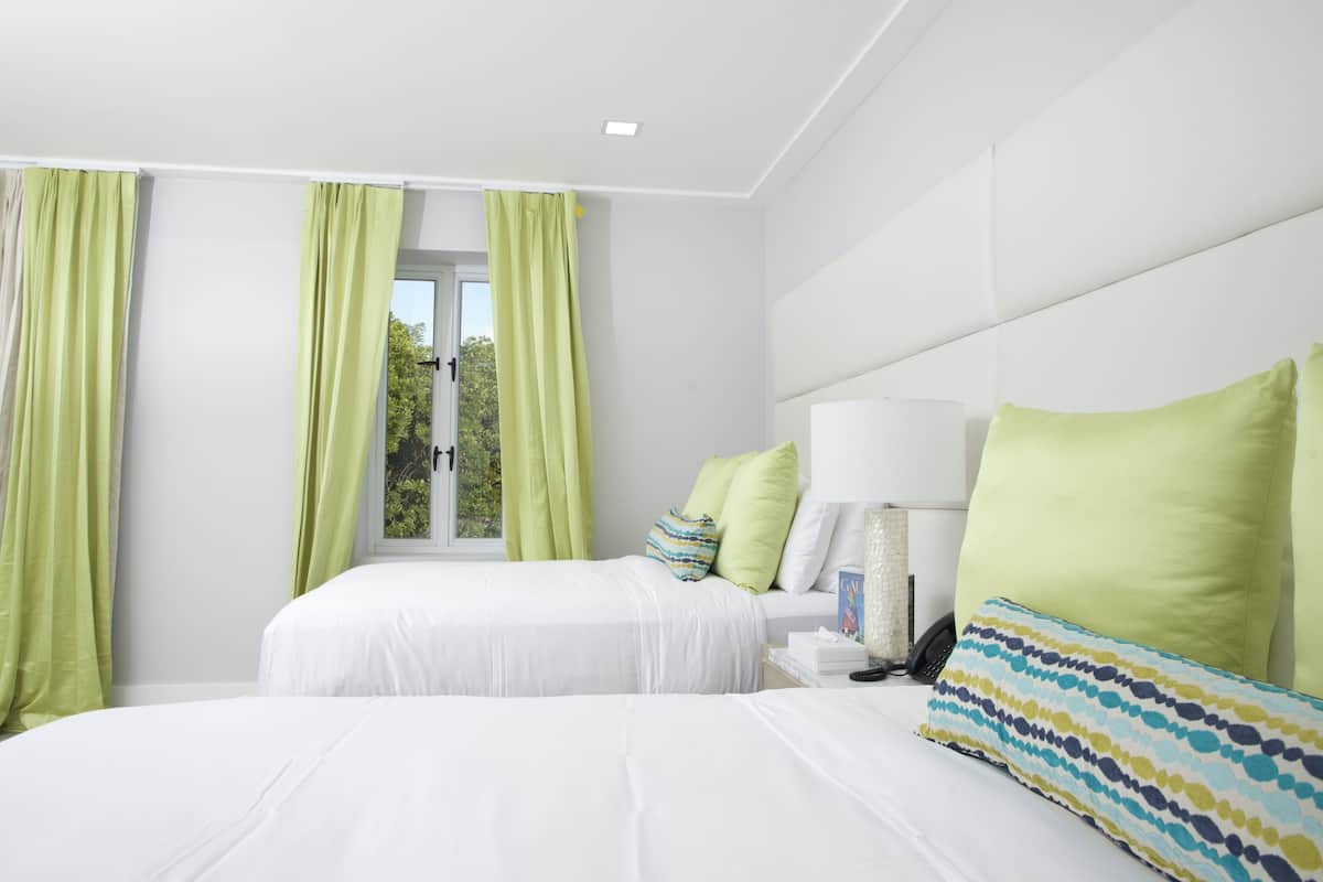 Habitación de hotel con dos camas, cortinas verdes, una mesita de noche con lámpara y vistas al exterior.