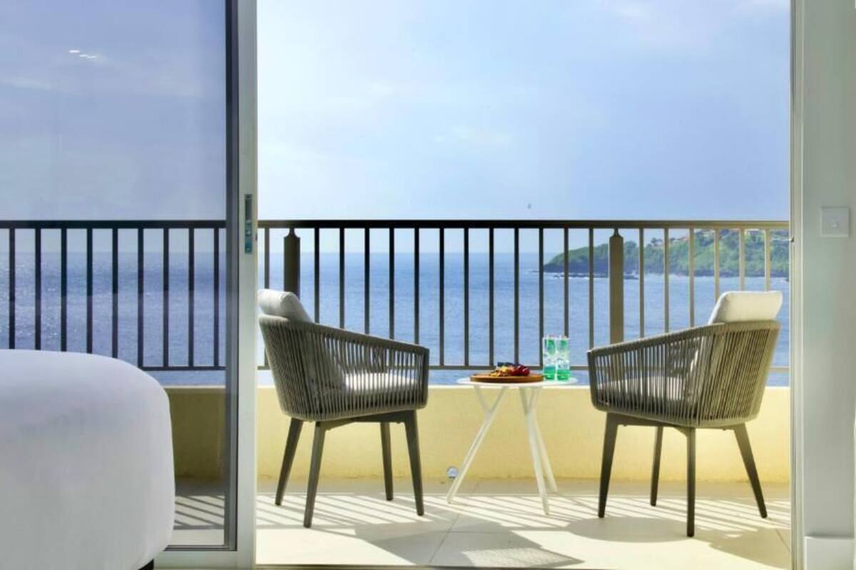 Un balcón con dos sillas de mimbre, una mesita con un plato de comida, y vistas al mar y una isla distante.