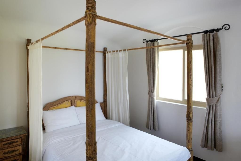 Un lit à baldaquin avec du linge de lit blanc, dans une pièce dotée d’une fenêtre et de rideaux.