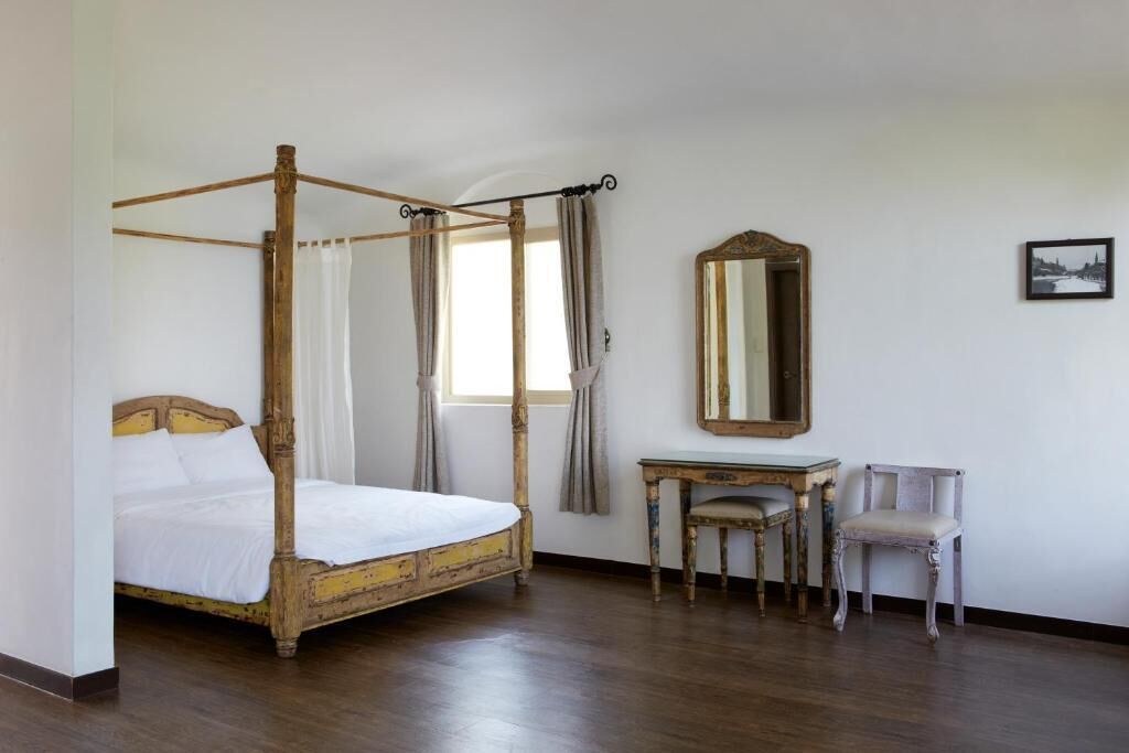 Un lit à baldaquin avec une structure en bois et du linge de lit blanc, une petite table avec une chaise, un miroir au mur et une fenêtre avec des rideaux.