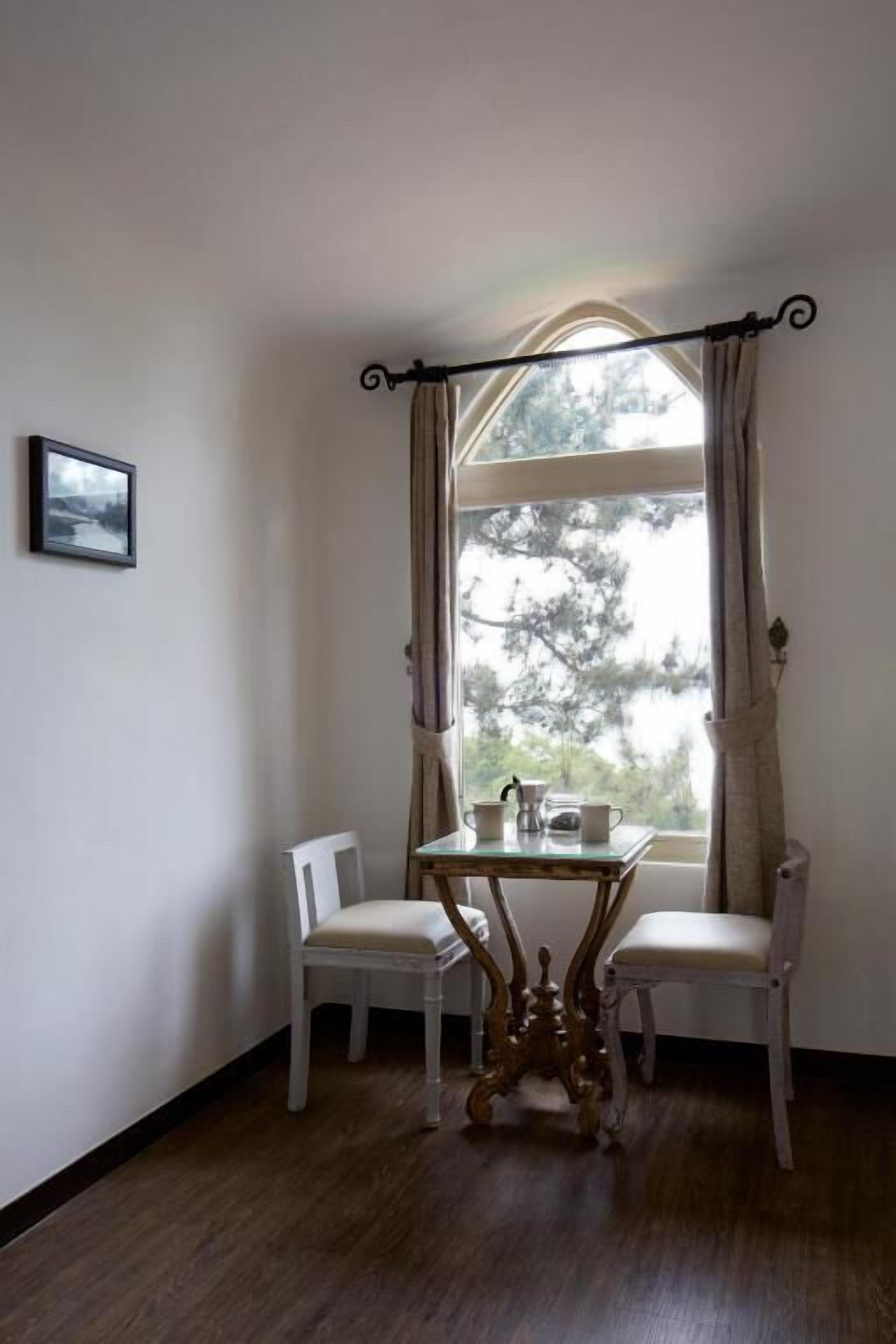 A corner room with a window, two chairs, a table with a teapot, and a framed picture on the wall.