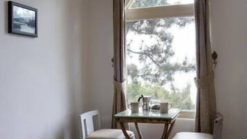 A corner room with a window, two chairs, a table with a teapot, and a framed picture on the wall.