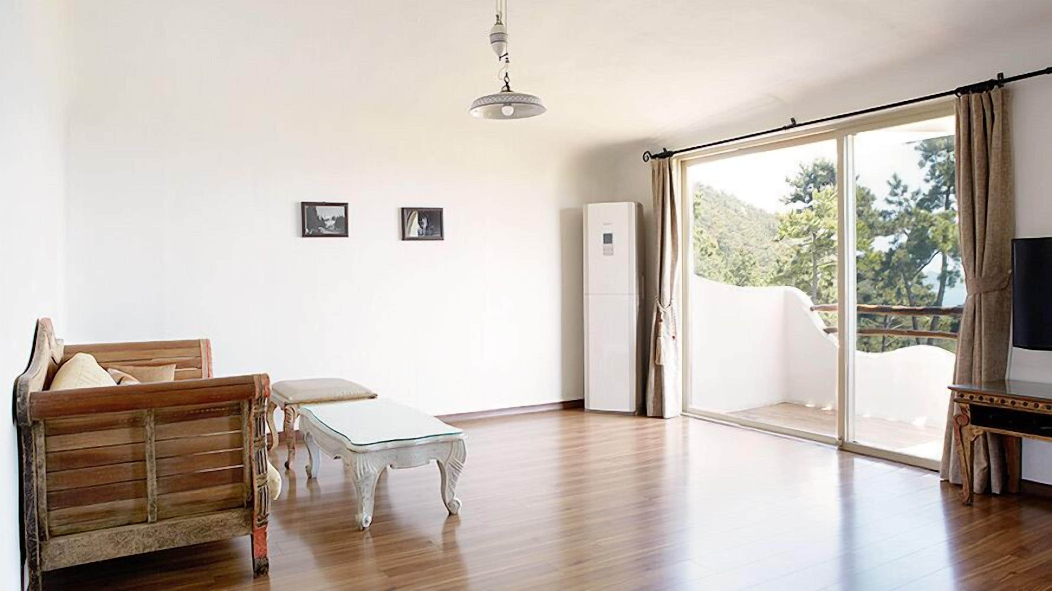 Uma sala de estar espaçosa com um sofá de madeira, uma mesa de centro de vidro e uma televisão sobre um móvel. Há uma porta de correr de vidro que dá para uma varanda com vista para o verde.