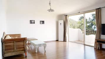 Uma sala de estar espaçosa com um sofá de madeira, uma mesa de centro de vidro e uma televisão sobre um móvel. Há uma porta de correr de vidro que dá para uma varanda com vista para o verde.