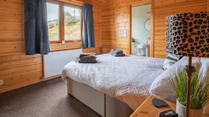 Une chambre à coucher dans une cabane en bois, avec un lit, une fenêtre avec des rideaux, une salle de bain visible à travers une porte ouverte, et une lampe à motifs léopard.