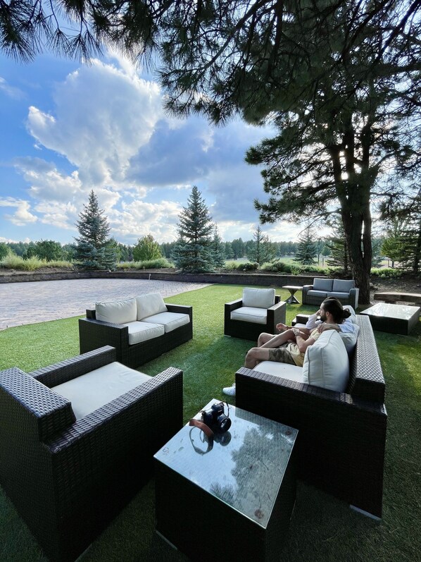 Un espacio al aire libre con mobiliario de mimbre y una mesa de cristal, con vistas a un cuerpo de agua y un parque.