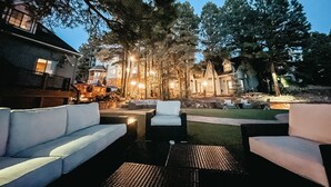 An outdoor seating area with sofas and a coffee table, surrounded by trees and a building with multiple windows.