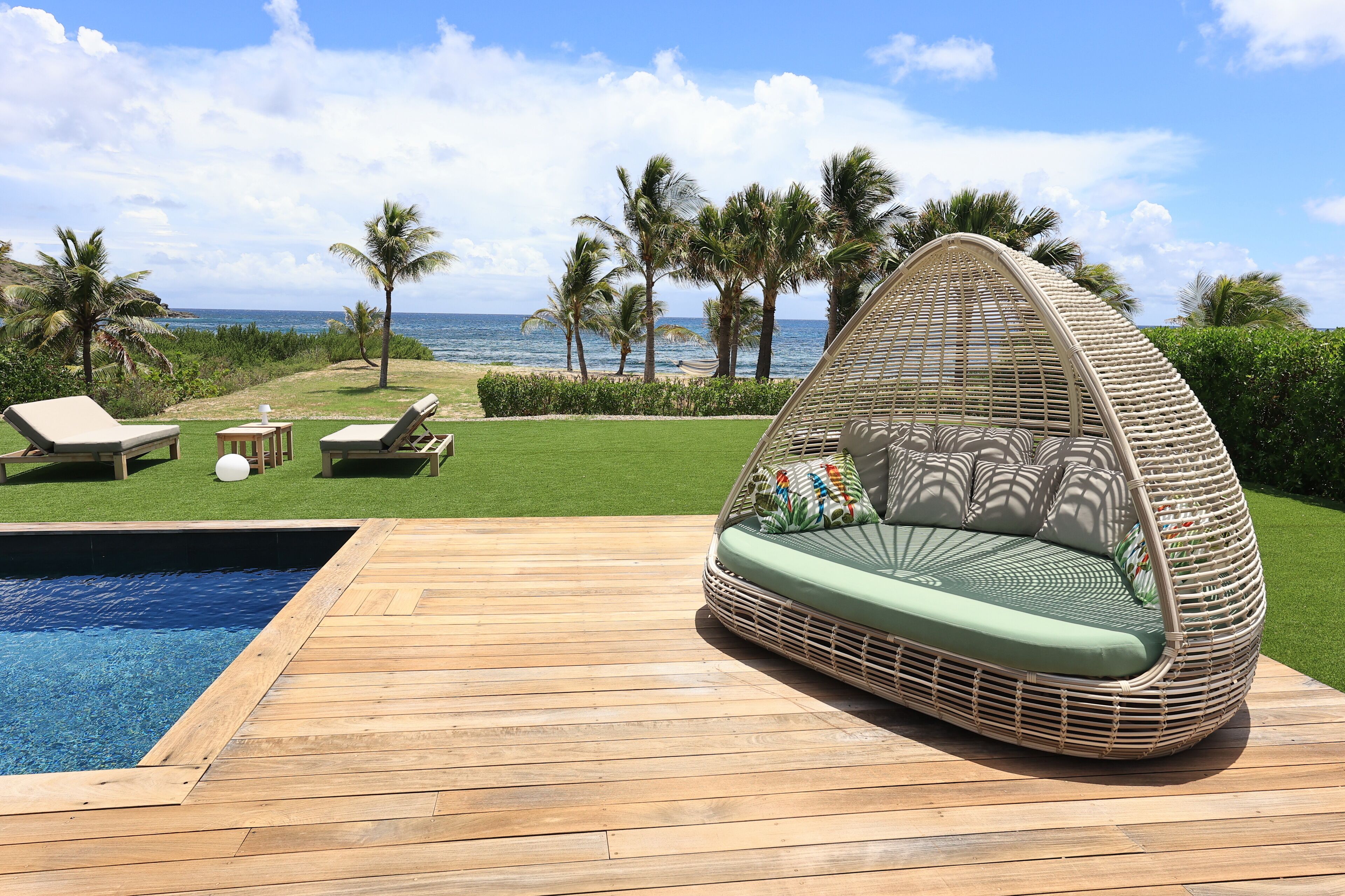 A wicker lounge chair with cushions by a pool, a wooden deck, and a view of the ocean and palm trees.