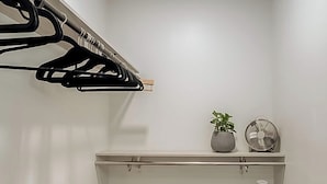 Een kleine, goed verlichte kamer met een houten droogrek, een zwarte dressoir, een klein plankje met een plant en een ventilator, en een kamerbreed tapijt met een patroon.