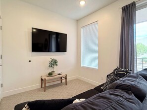 A living room with a flat-screen TV mounted on the wall, a small wooden bench with a plant, a large black sofa, and a window with blinds.