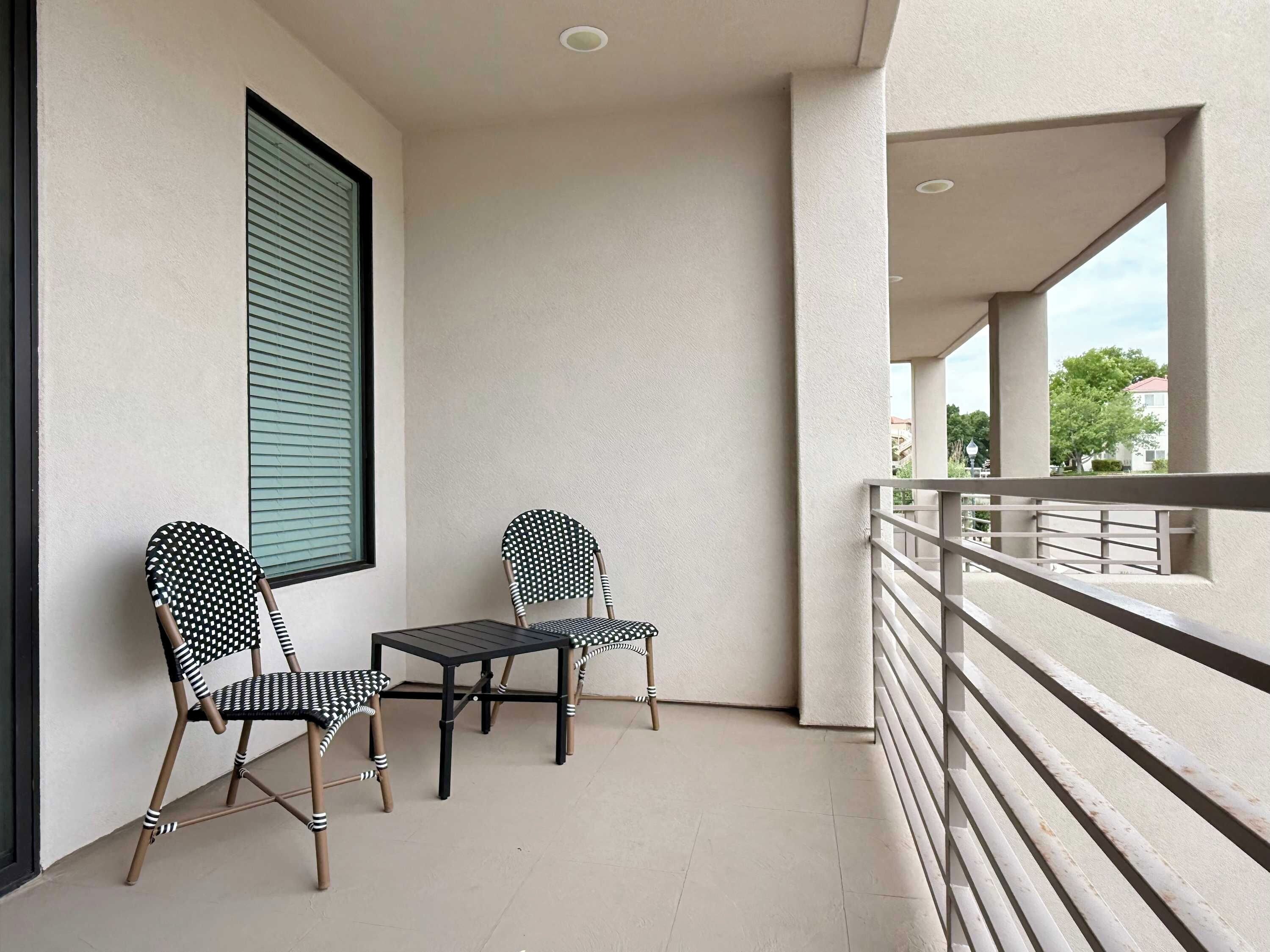 Ein Balkon mit zwei Stühlen und einem Tisch, ein Fenster mit Jalousien und Blick auf die Straße.