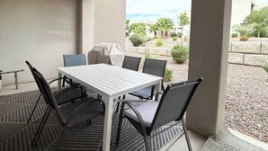 An outdoor patio with a table and chairs, a grill, and a view of a landscaped area with trees and a building.