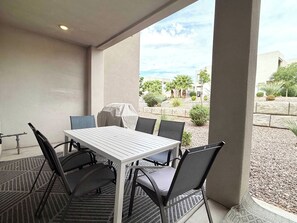 An outdoor patio with a table and chairs, a grill, and a view of a landscaped area with trees and a building.