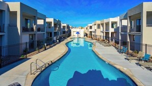 A swimming pool surrounded by modern apartment buildings with balconies, a clear sky, and lounge chairs along the poolside.