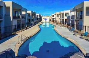 A swimming pool surrounded by modern apartment buildings with balconies, a clear sky, and lounge chairs along the poolside.