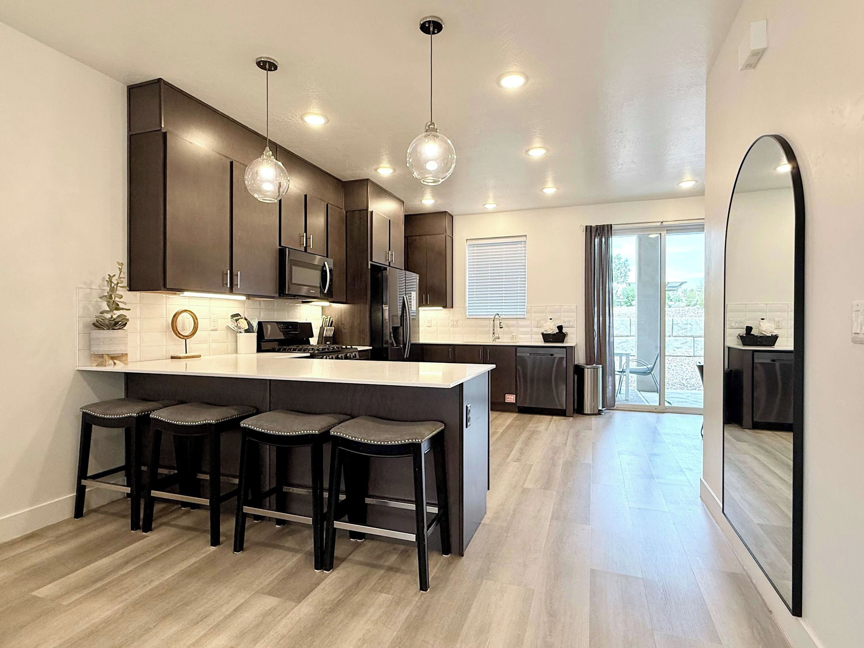 A modern kitchen with a central island, bar stools, stainless steel appliances, pendant lighting, and a view to the outside.