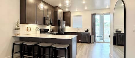 A modern kitchen with a central island, bar stools, stainless steel appliances, pendant lighting, and a view to the outside.