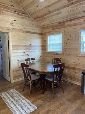 Una habitación con paneles de madera, una mesa de comedor y sillas, una ventana con persianas y una puerta que conduce a otra habitación.