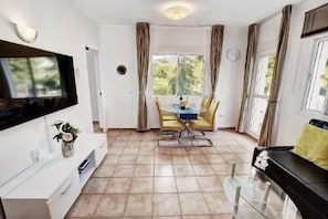 A modern room with a flat-screen TV mounted on the wall, a dining area with a table and chairs, and large windows with curtains.