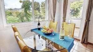 Un comedor con una mesa de vidrio, sillas amarillas, una botella de vino, un cuenco de fruta y un vaso de agua.