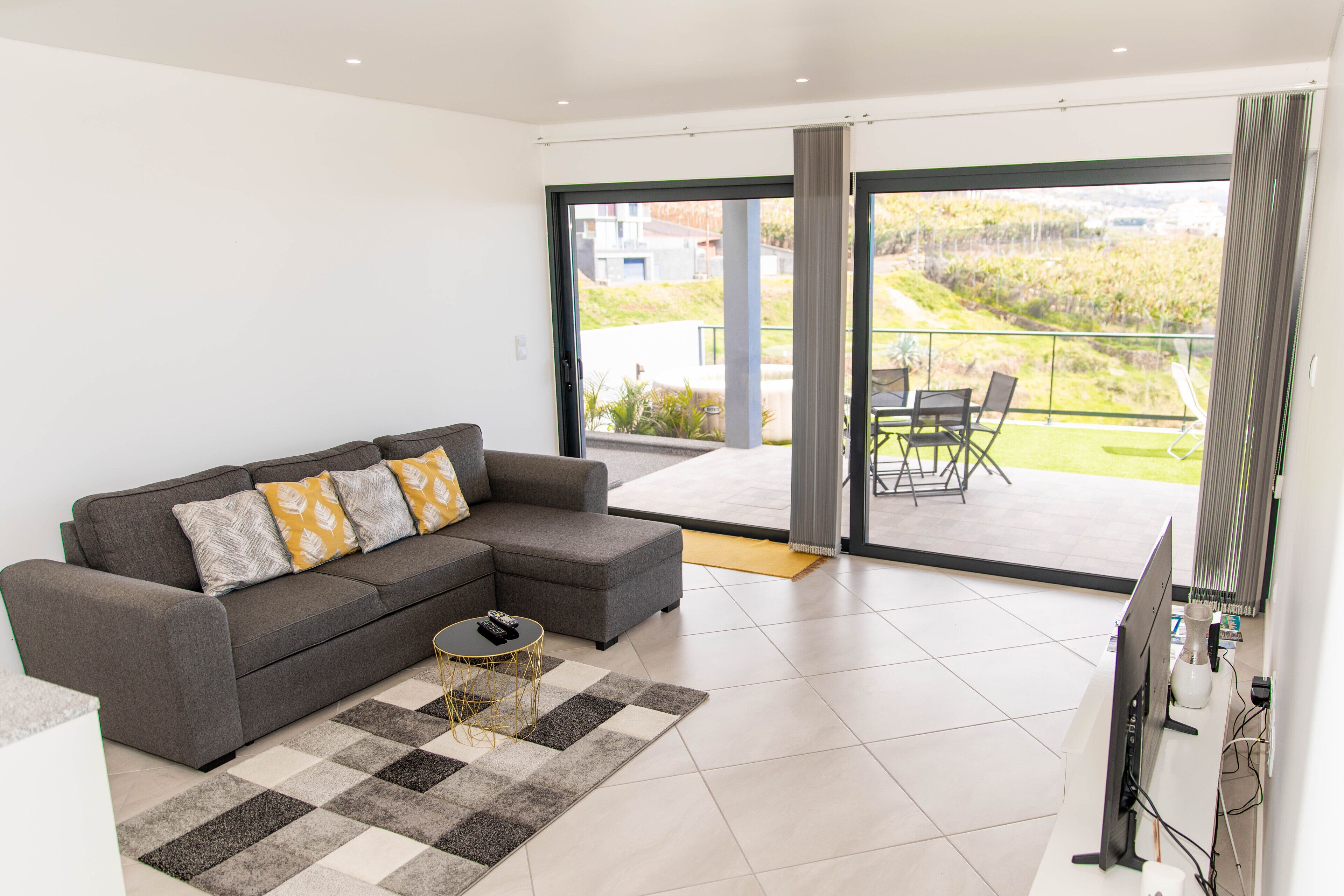 Een moderne woonkamer met een grijze modulaire bank, een klein rond salontafeltje en een flatscreen televisie. De kamer heeft een grote schuifdeur die leidt naar een balkon met uitzicht op groen.