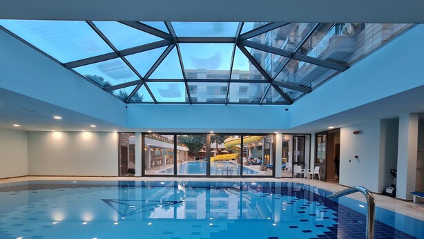 A modern indoor swimming pool with a glass ceiling, clear pool tiles, and a view of an outdoor area with a slide. - 2-Bedroom, Sleeps 4, Free Parking (Vlore)
