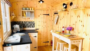 A compact kitchen with wooden walls, a black countertop, a sink, a stove, and a bar area with stools. - Woodhead Shepherds Hut - Sleeps 2 - Parking (Perth)