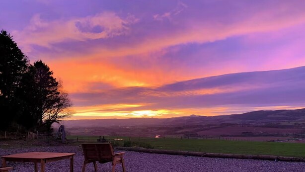 A sunset view from a gravel patio with two wooden chairs and a table, overlooking a rural landscape with fields and hills. - Woodhead Shepherds Hut - Sleeps 2 - Parking (Perth)