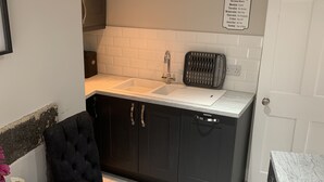 Une cuisine de taille réduite, dotée de meubles de rangement sombres, d’un plan de travail blanc et d’un évier. Un panneau indiquant les heures d’ouverture de la cuisine est placé au-dessus de l’évier.