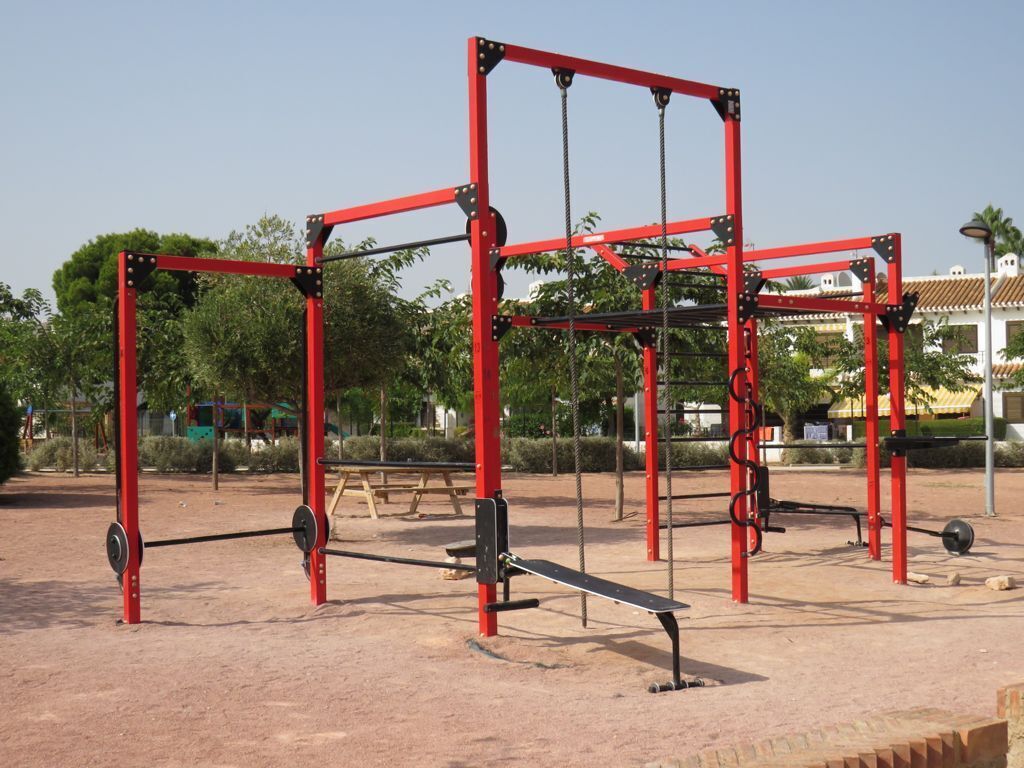 Une structure métallique rouge, dotée d’une barre de traction et de poids, installée dans un espace extérieur, avec des arbres et des bâtiments en arrière-plan.