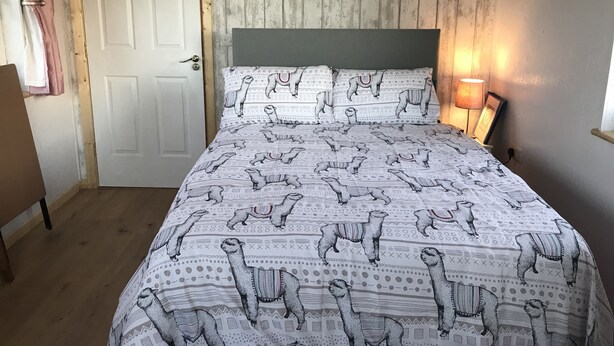 A bedroom with a patterned bedspread, a wooden door, and a bedside lamp. - The Lazy Llama Shepherd Hut & Sauna (Ballyshannon)