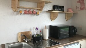 A kitchen with wooden shelves holding mugs and boxes, a microwave, and a sink with a cutting board. - The Lazy Llama Shepherd Hut & Sauna (Ballyshannon)