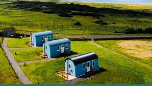 A rural landscape with three blue cabins, a stone structure, and a fenced area. - The Lazy Llama Shepherd Hut & Sauna (Ballyshannon)