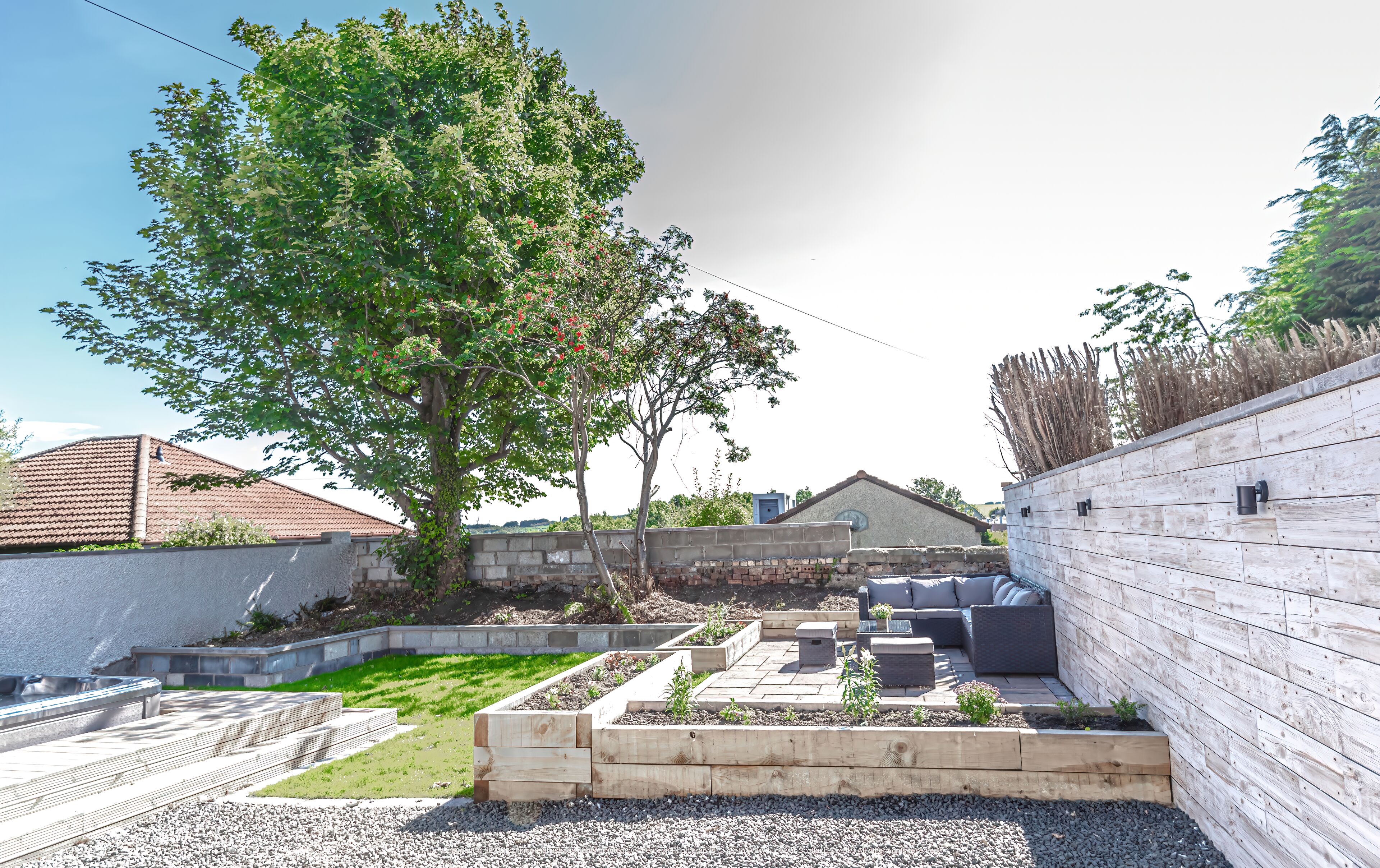 Un patio trasero con terraza de madera, jardines elevados y mobiliario exterior.