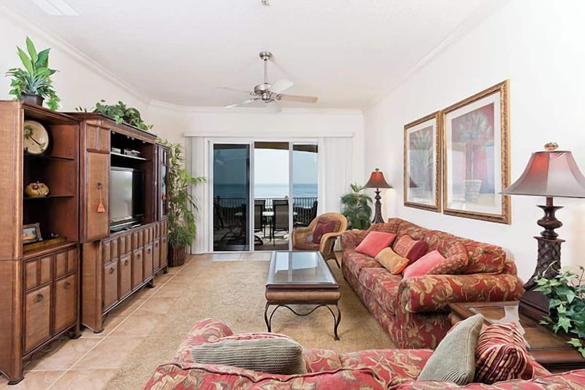 A living room with a sofa, coffee table, and TV stand. There is a sliding glass door leading to a balcony.