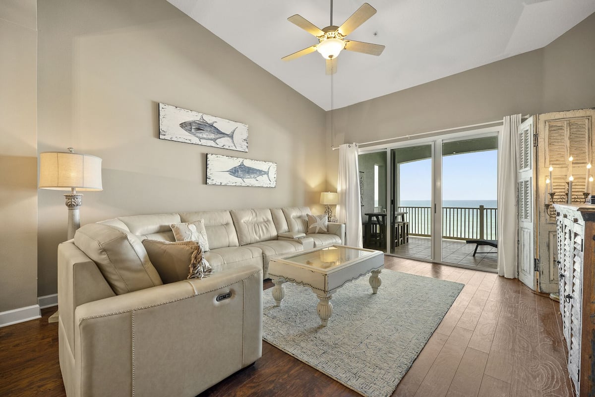 A spacious living room with a beige sofa, a glass coffee table, and a ceiling fan. There are two framed fish paintings on the wall and a view of the ocean through the sliding glass doors.