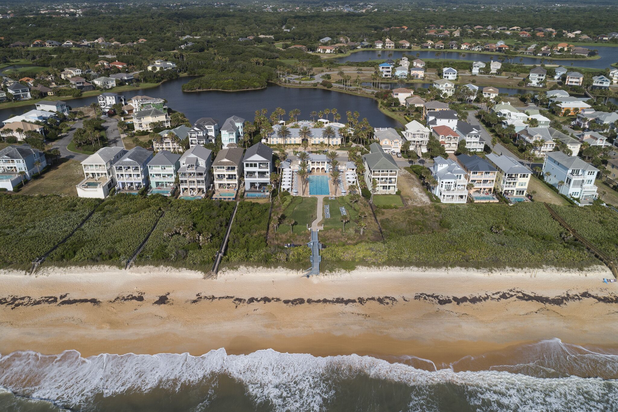 An aerial view of a coastal residential area with houses, a beach, and a lake.