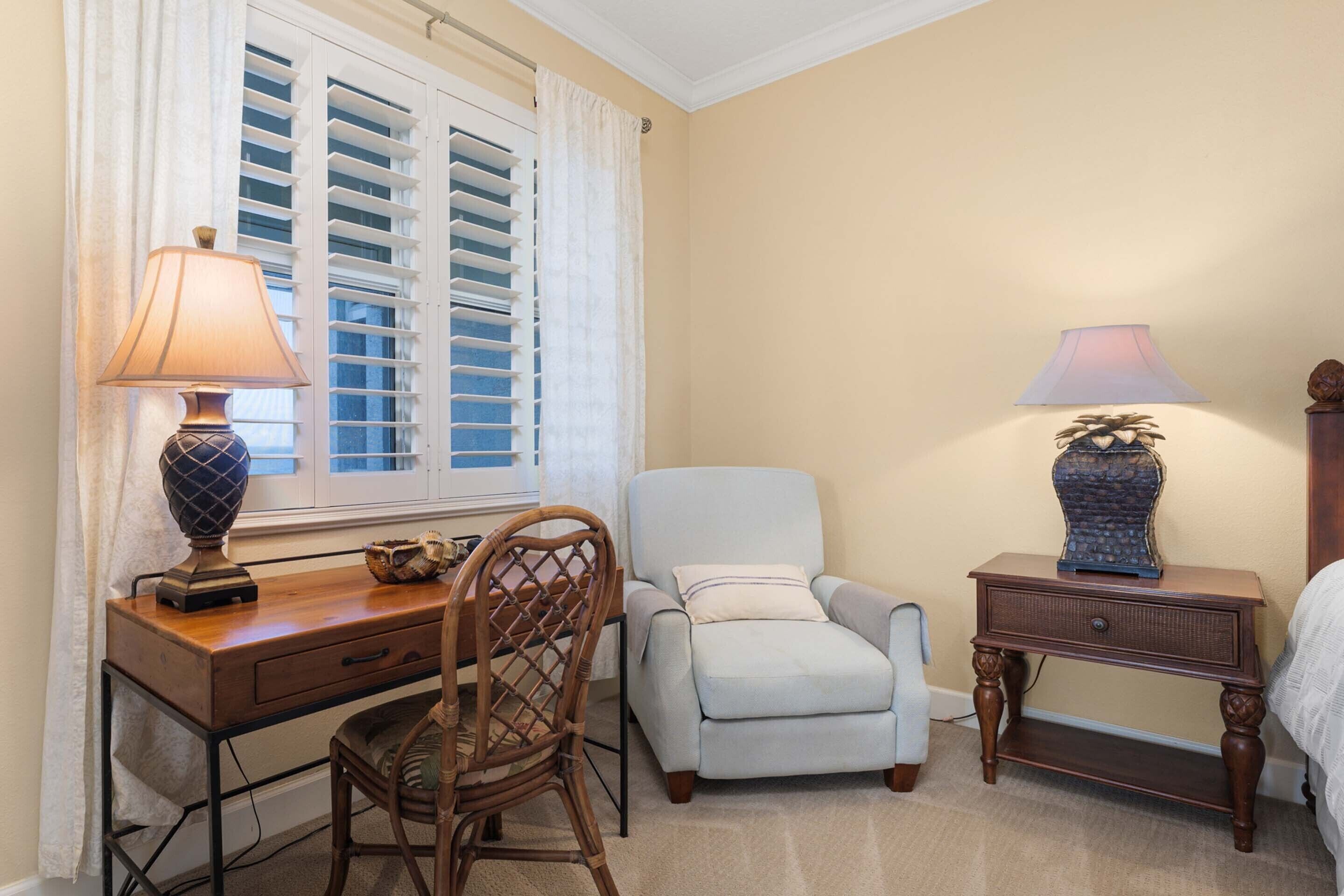A room with a desk, chair, armchair, and bedside table with lamps.