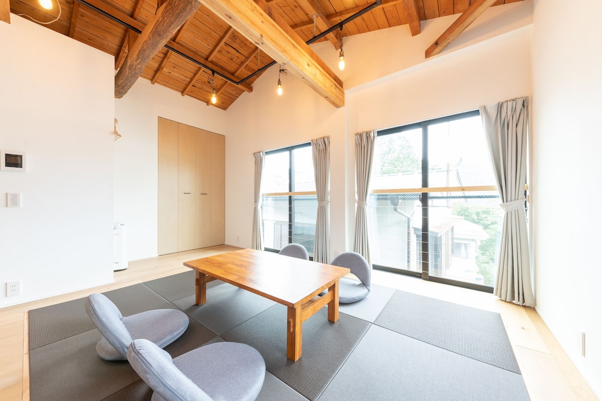 Una habitación amplia con una mesa y sillas de madera, ventanas grandes con cortinas y un techo alto con vigas a la vista.