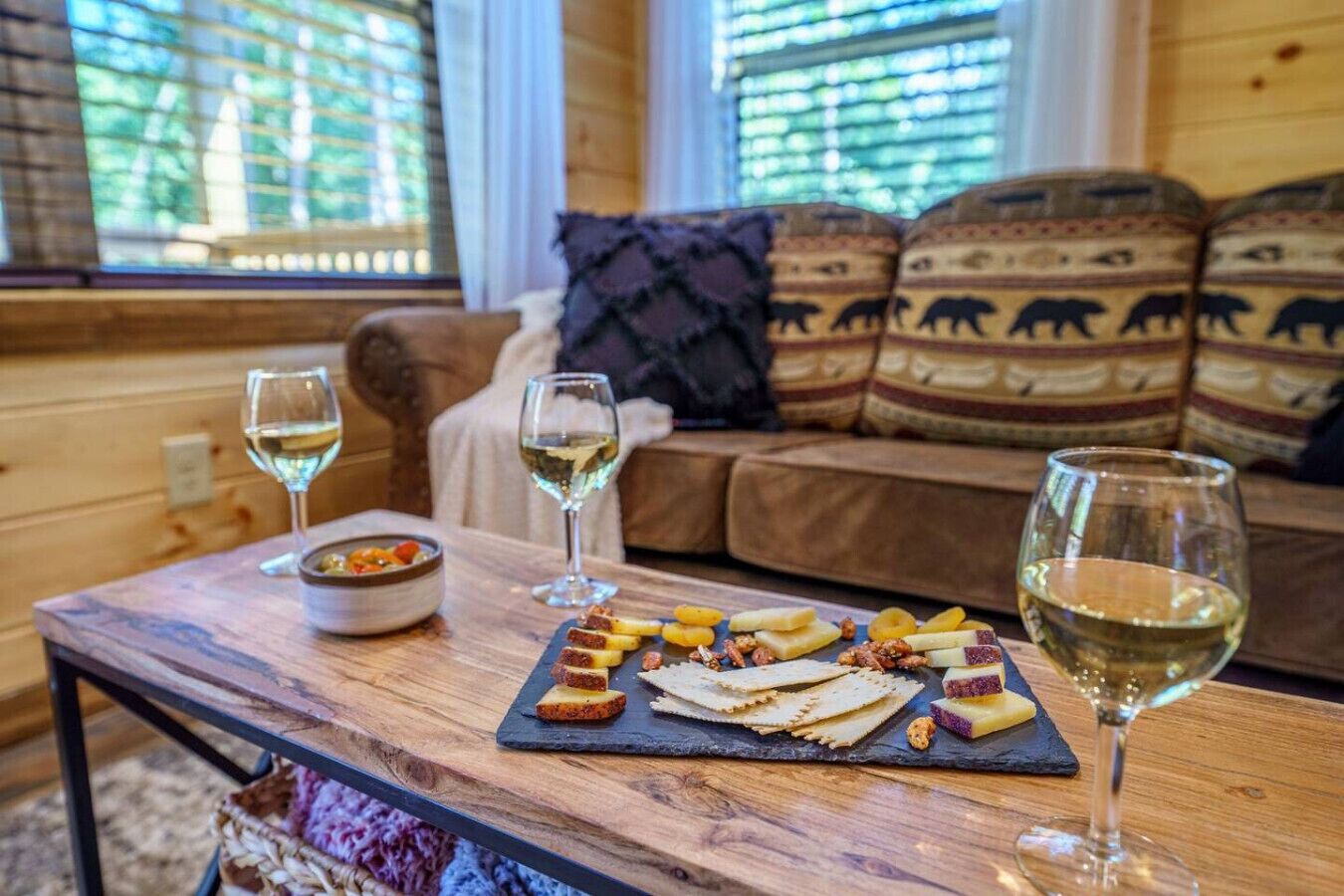 Une pièce chaleureuse avec une table en bois où sont disposés des en-cas et des verres à vin, un canapé agrémenté de coussins à motifs et une fenêtre avec des stores.