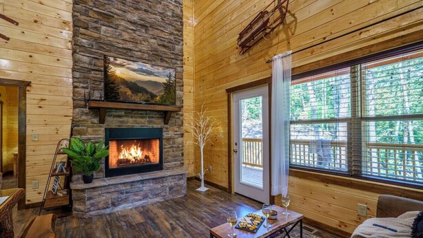 A cozy cabin interior with a stone fireplace, wooden walls, a large window, and a dining area with a table set for two. - Great Location! Pigeon Forge, Pool Table/Hot Tub & Game Room, Quiet Neighborhood (Sevierville)