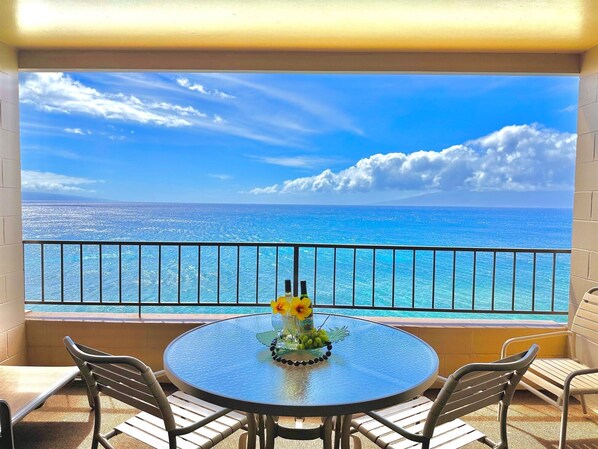 A round table with two chairs, a glass top, and a view of the ocean through a balcony railing.