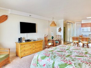 A bedroom with a bed, dresser, television, dining table, chairs, and a kitchen area.