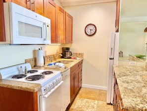 A kitchen with wooden cabinets, a white stove, a microwave, a clock, and a coffee maker. - Junior Suite Oceanfront #907 (Lahaina)