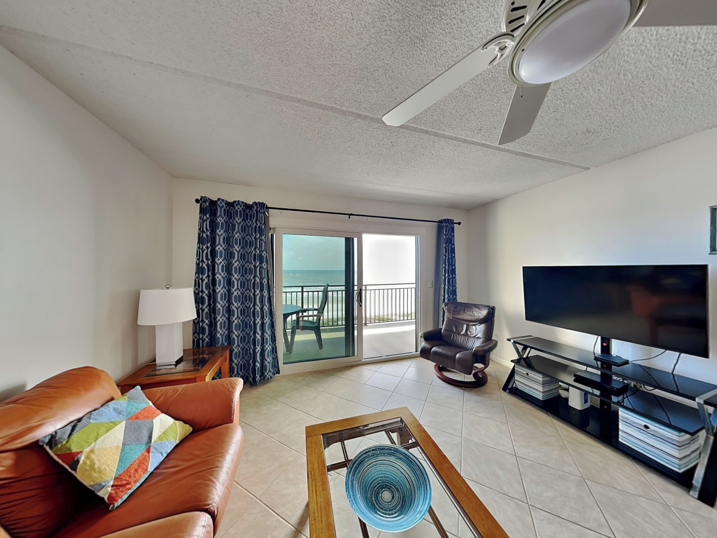Una sala de estar con un sofá de cuero marrón, una cortina azul estampada, un televisor y un balcón con vista al mar.